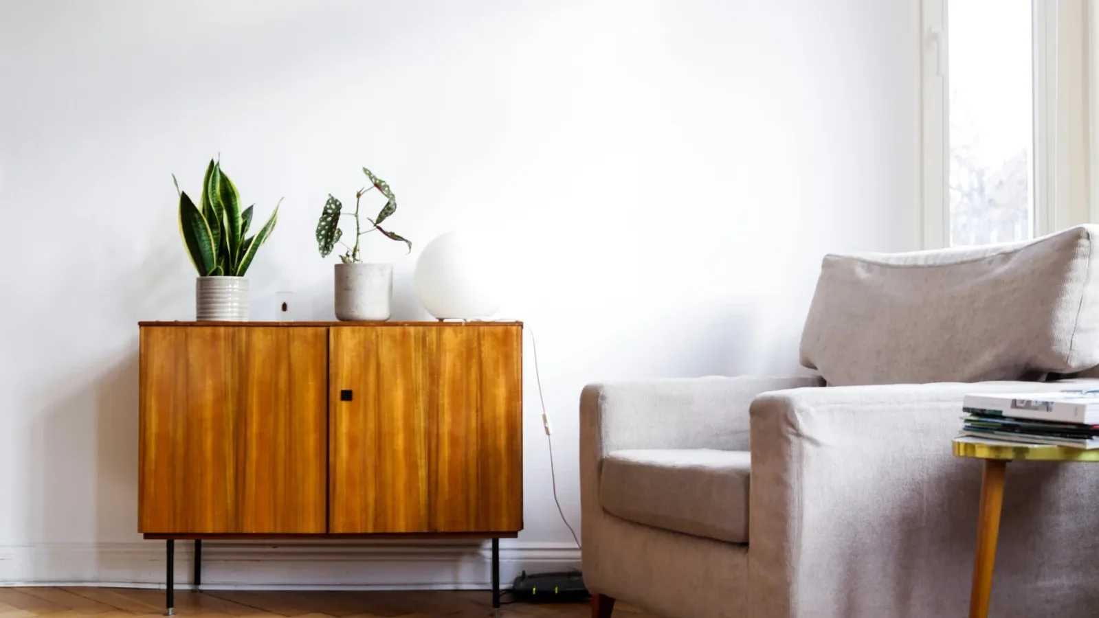 Bright minimalist living room with beige armchair, mid-century wooden cabinet, potted plants, and herringbone wood flooring.