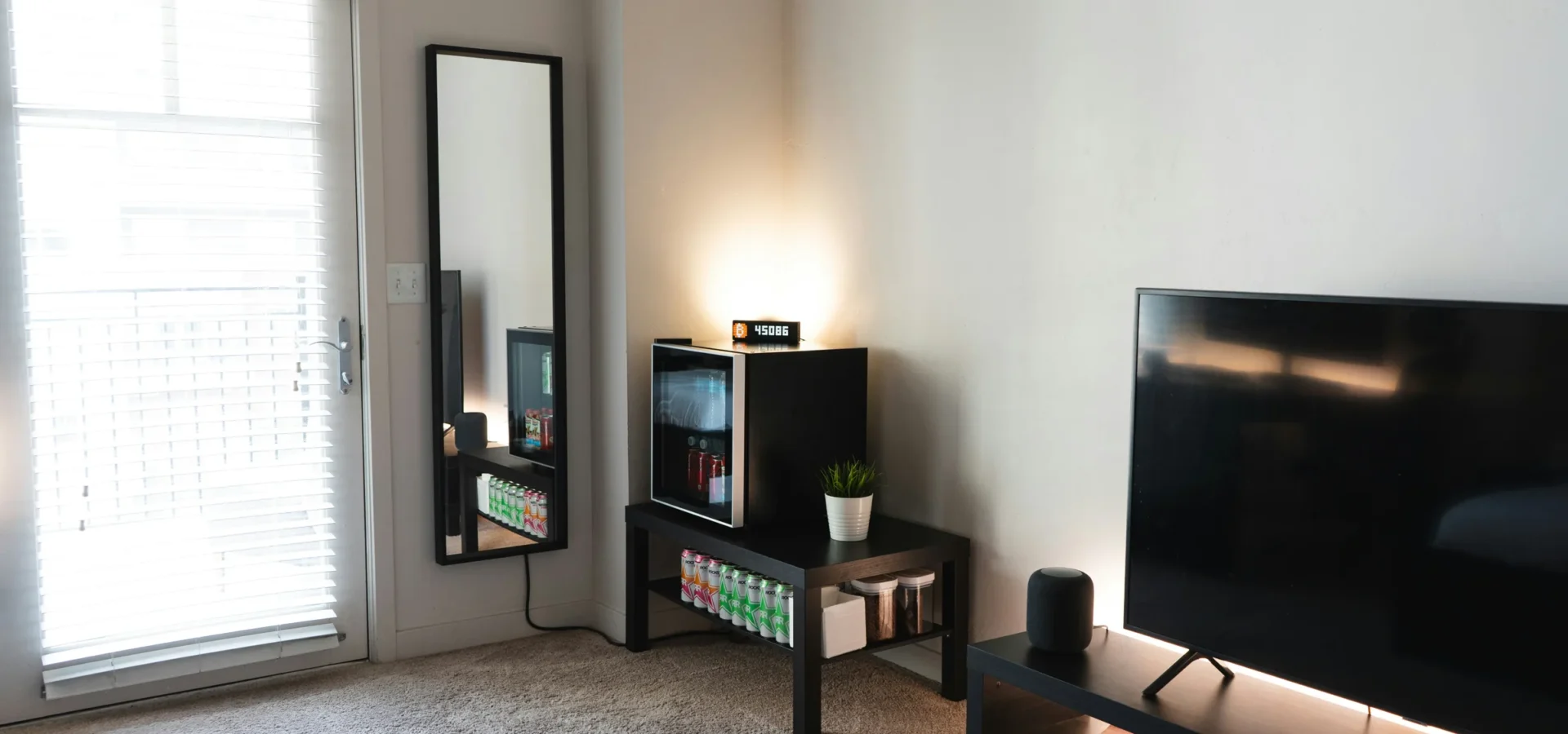 Modern living room with TV, beverage cooler, mirror, potted plant, and furniture on gray carpet near glass door.