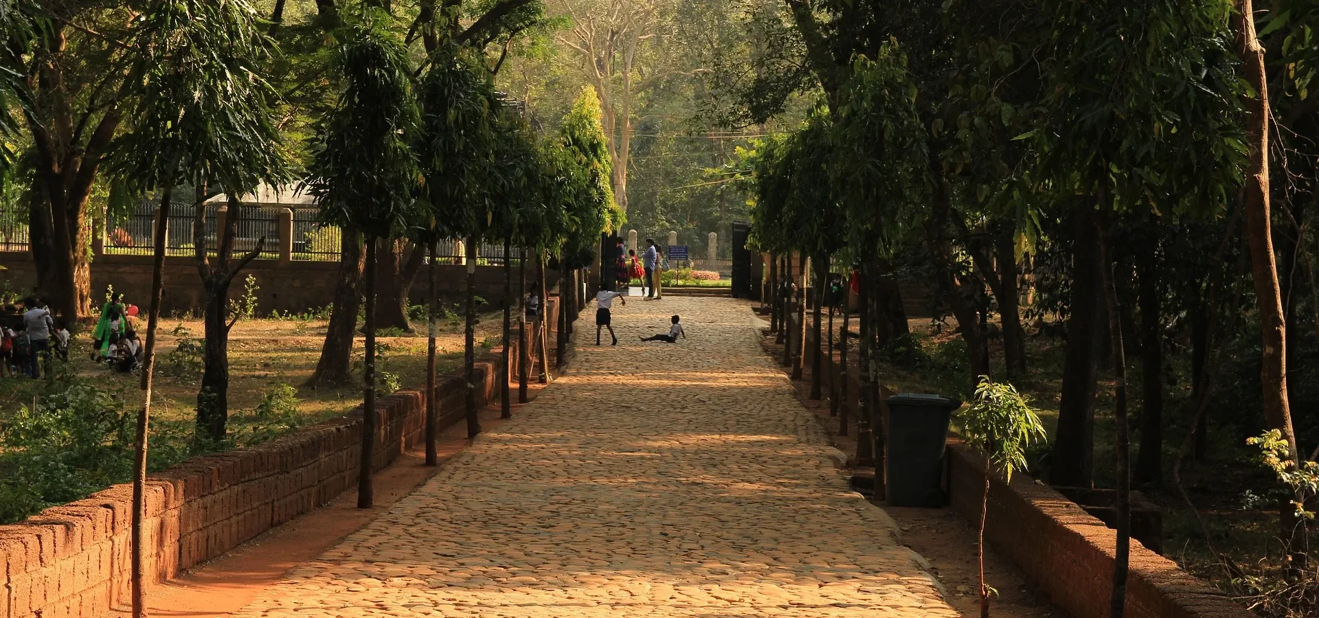 Tree-lined sandy pathway in a park with visitors walking and sitting, bordered by brick walls and lush green foliage.