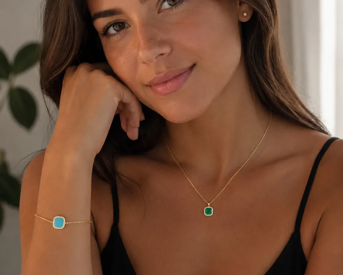 Woman wearing black top and gold jewelry with turquoise and emerald gemstone pendant and bracelet, posing indoors.