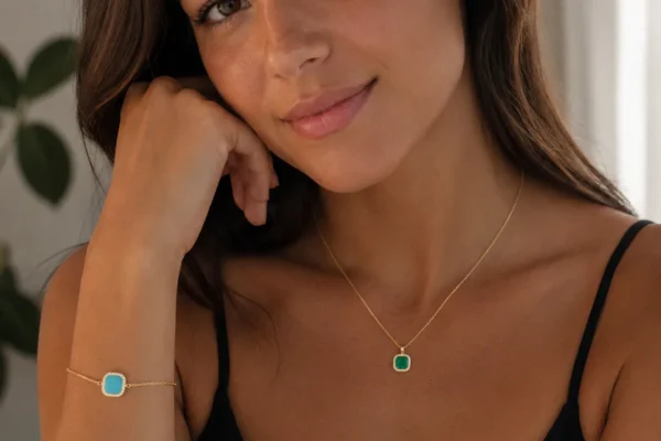 Woman wearing black top with turquoise and emerald gemstone jewelry, smiling at camera in bright indoor setting with plant background.