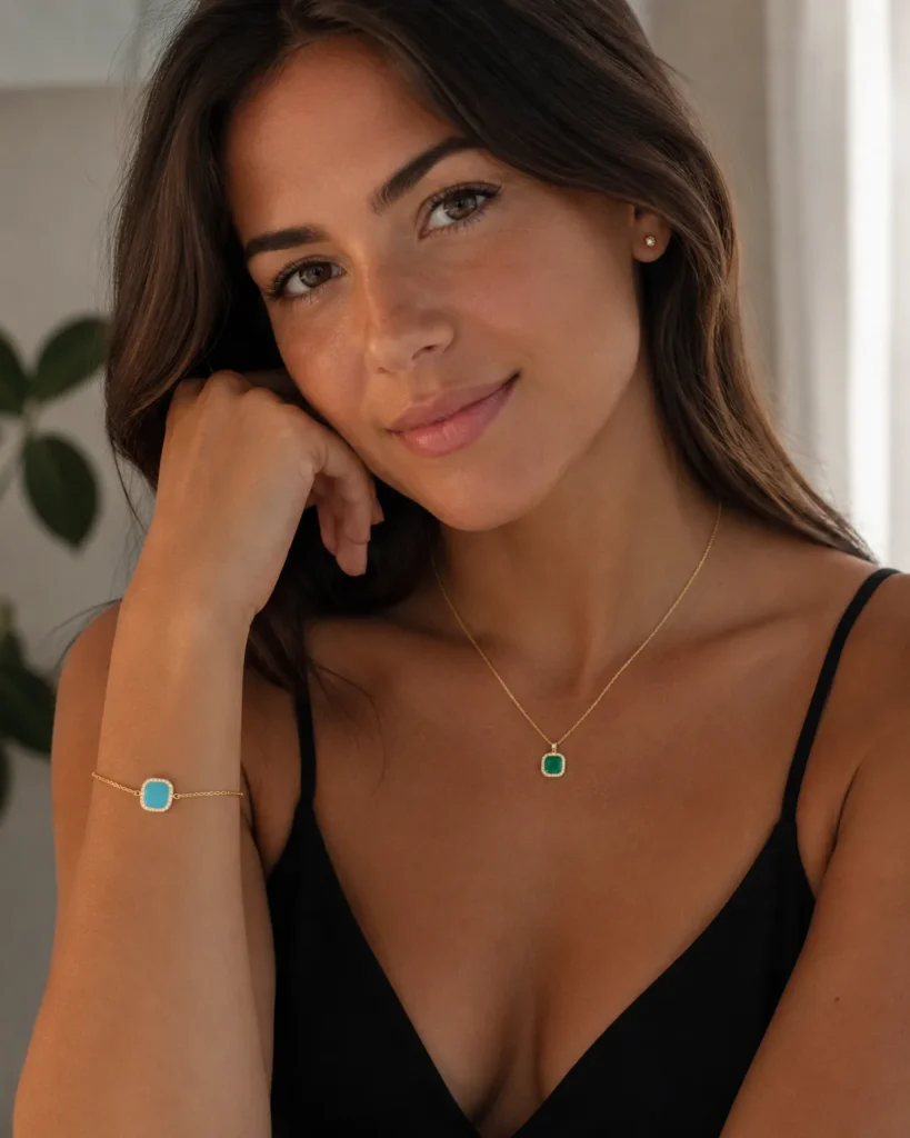 Woman wearing black top with turquoise and emerald gemstone jewelry, smiling at camera in bright indoor setting with plant background.